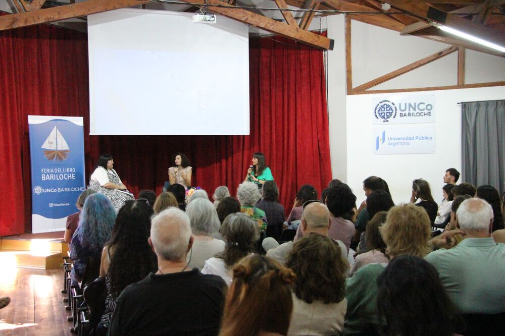 Tres mujeres conversan con micrófono en el escenario del salón del CRUB, frente a un público sentado que llena la sala.