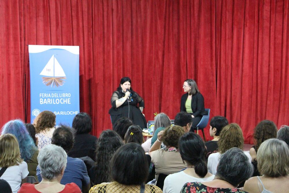 Dos mujeres charlan con micrófono en el escenario, junto al banner de la Feria del Libro Bariloche y frente al público sentado.